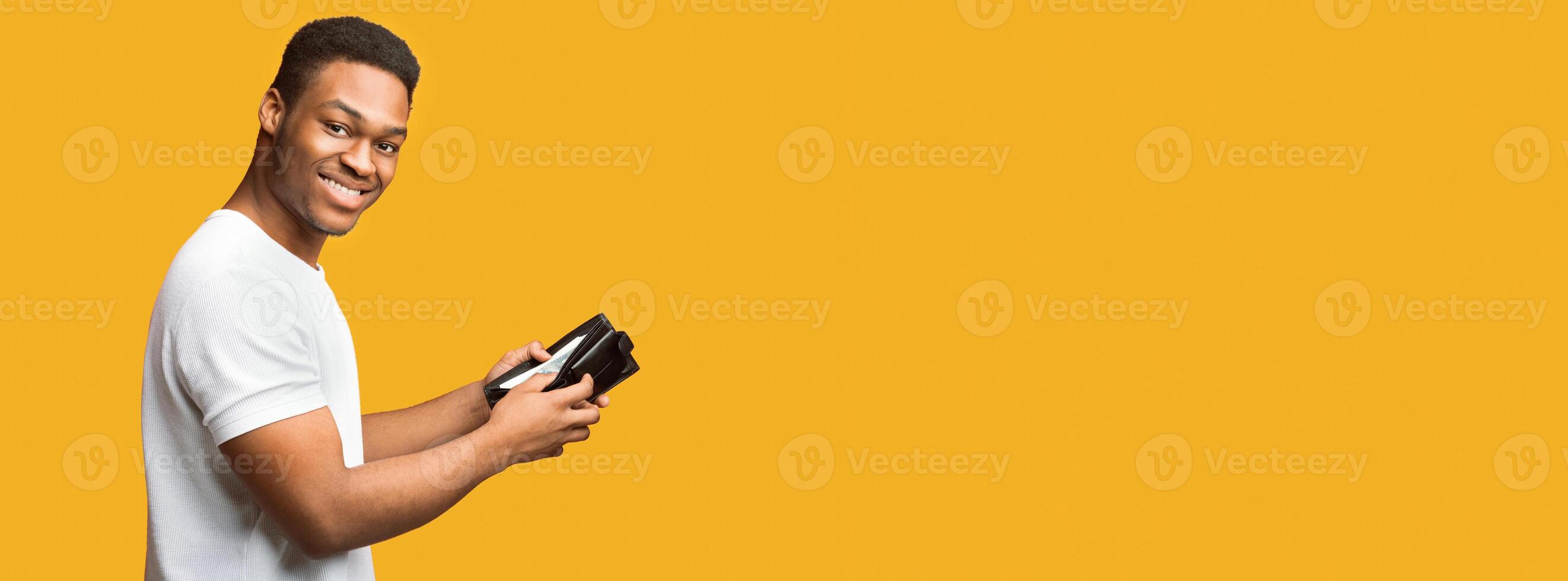 Money Saving. Smiling black man showing wallet full of dollars over yellow studio wall, panorama, copyspace photo