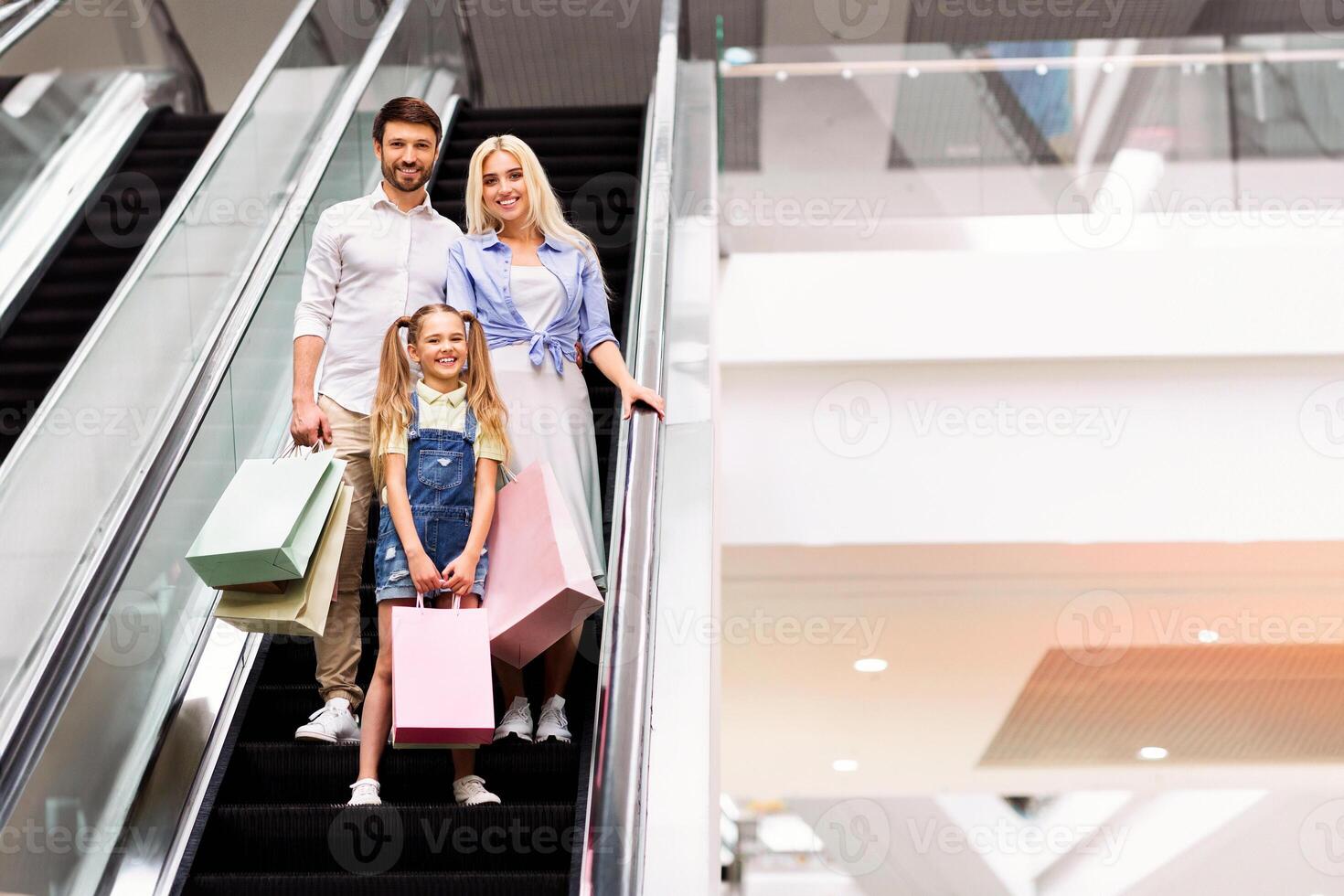 A happy family, a father, mother, and young daughter, are riding up an escalator in a modern shopping mall. They are all smiling and holding shopping bags, copy space photo