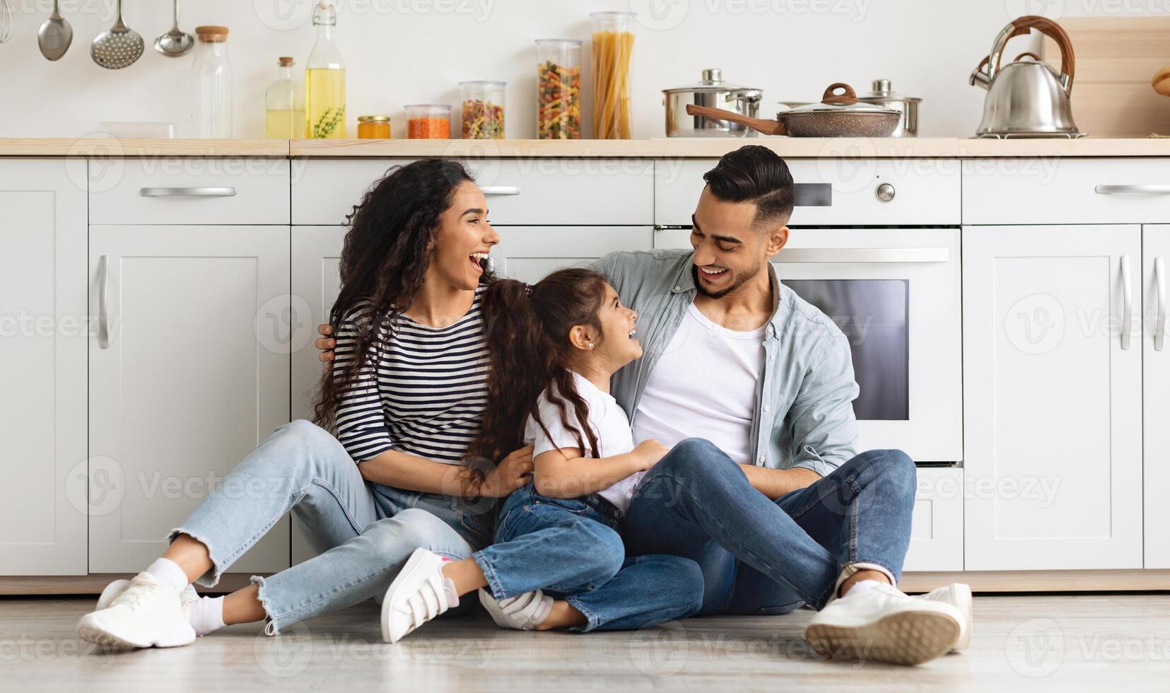 Happy arab family spending weekend together at home, cheerful middle-eastern young father, mother and cute little girl daughter sitting on tile floor at white kitchen, having conversation, copy space photo