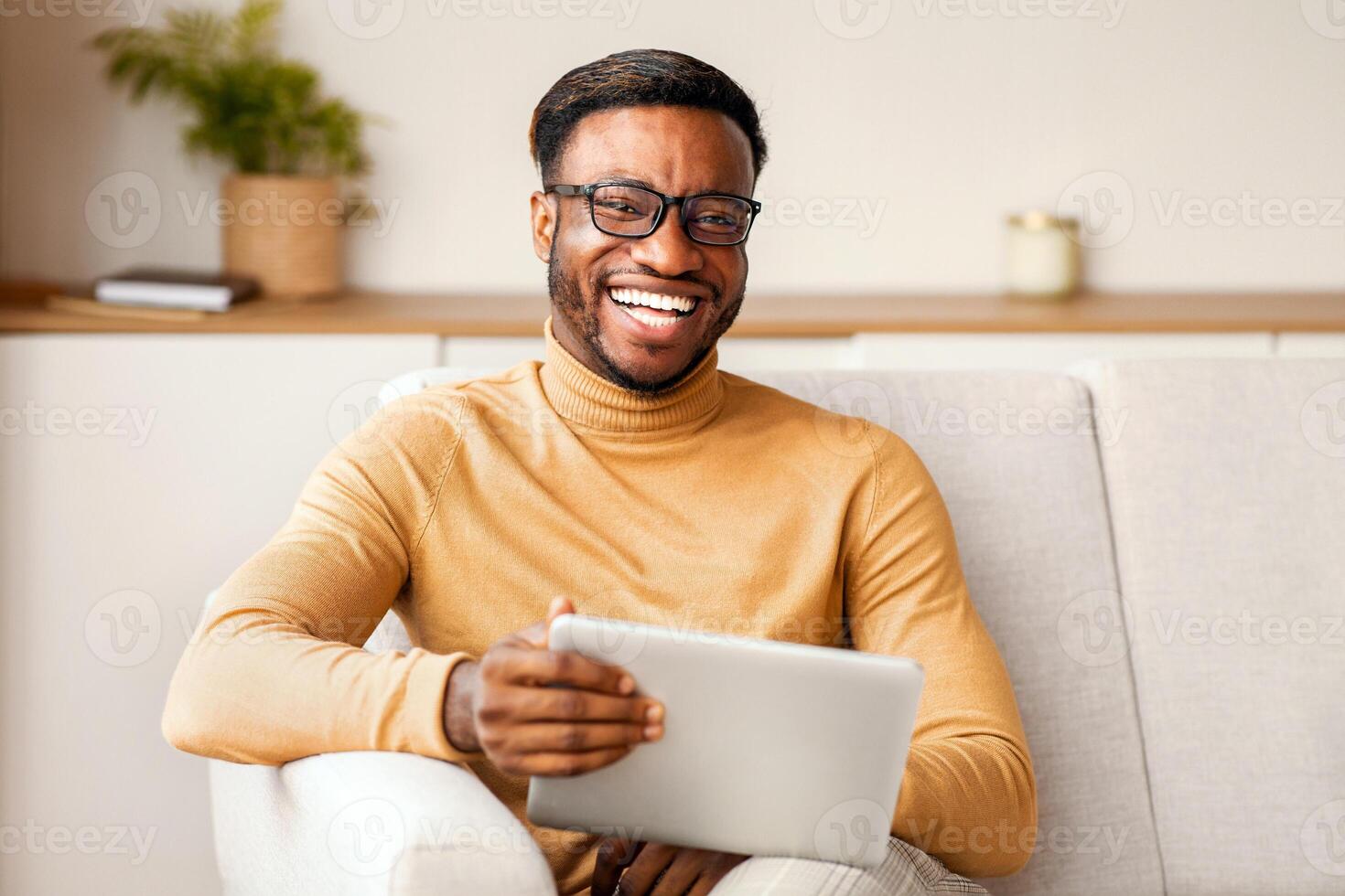 Positive Afro Man Holding Digital Tablet Laughing Looking At Camera Sitting On Couch At Home. Selective Focus photo