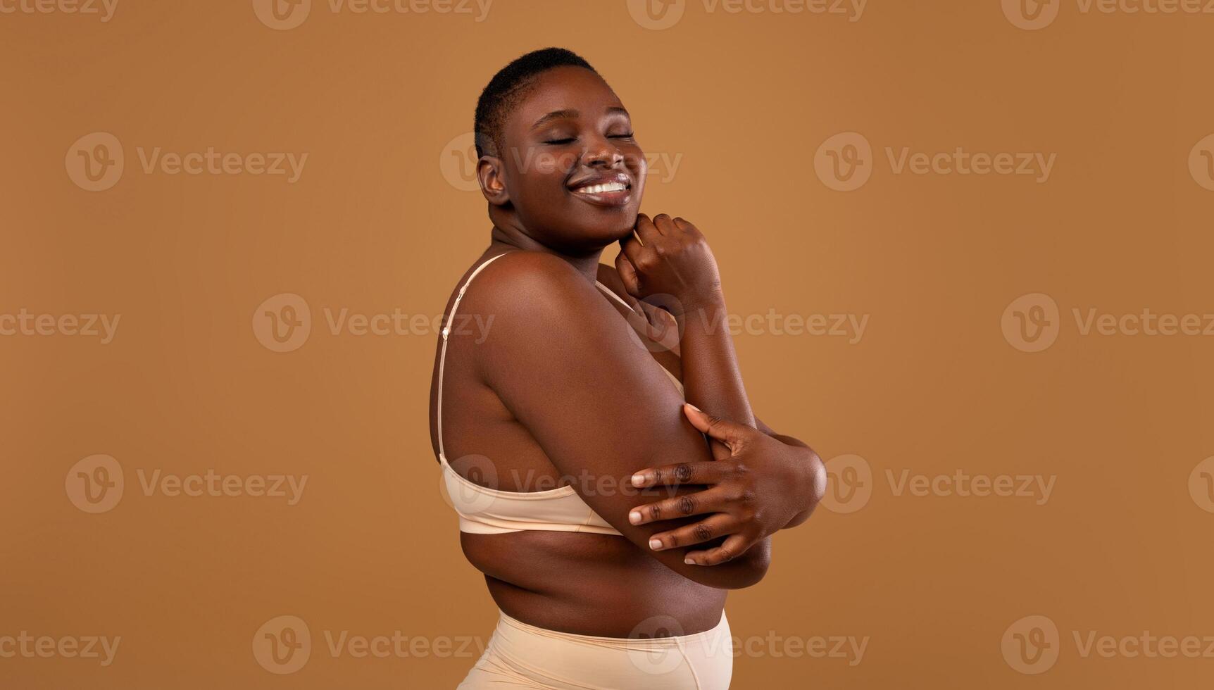 Self Acceptance, Love And Care. Portrair of half-turned smiling curvy African American woman with perfect skin and closed eyes embracing herself isolated on brown studio background, free copy space photo