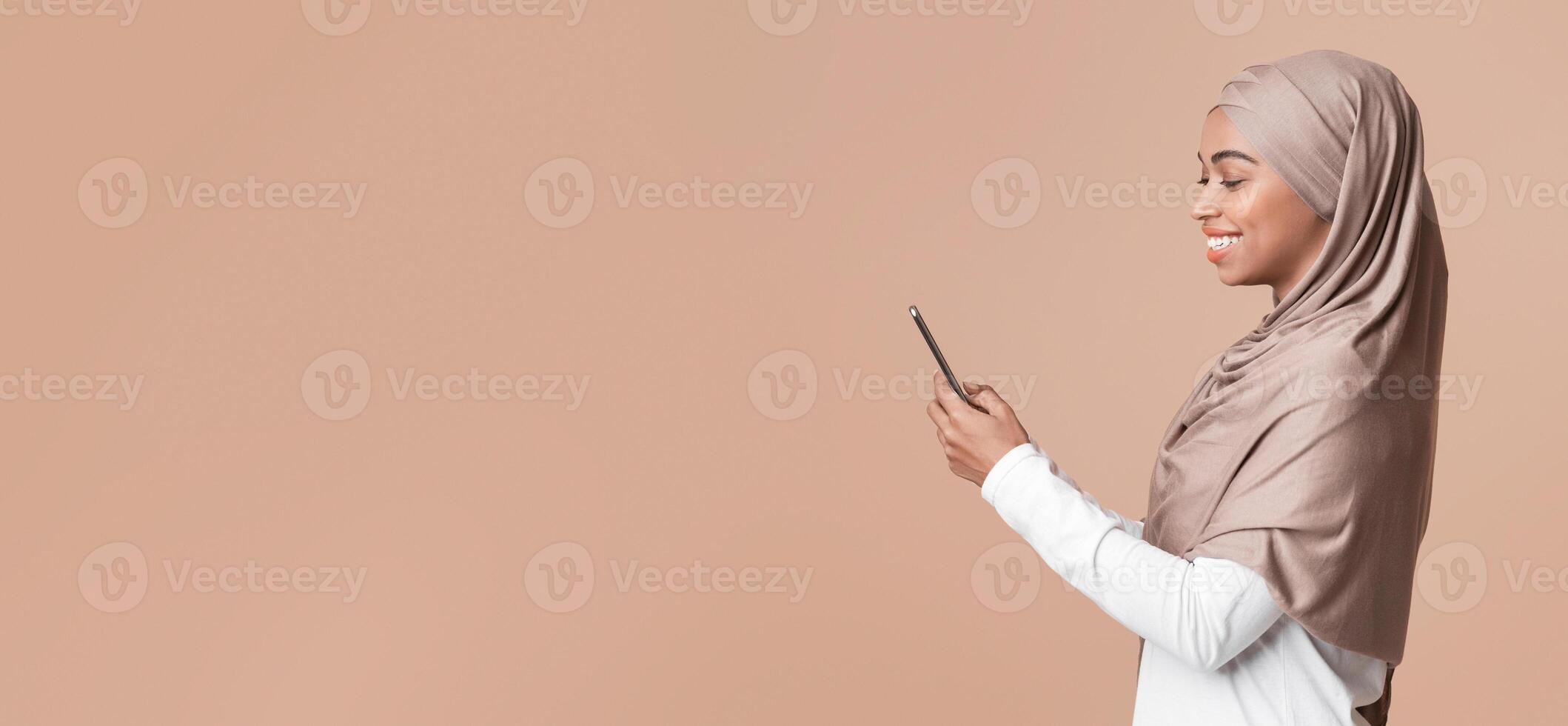 Profile portrait of cheerful black girl in hijab using smartphone, texting or browsing social media, yellow studio background with empty space photo