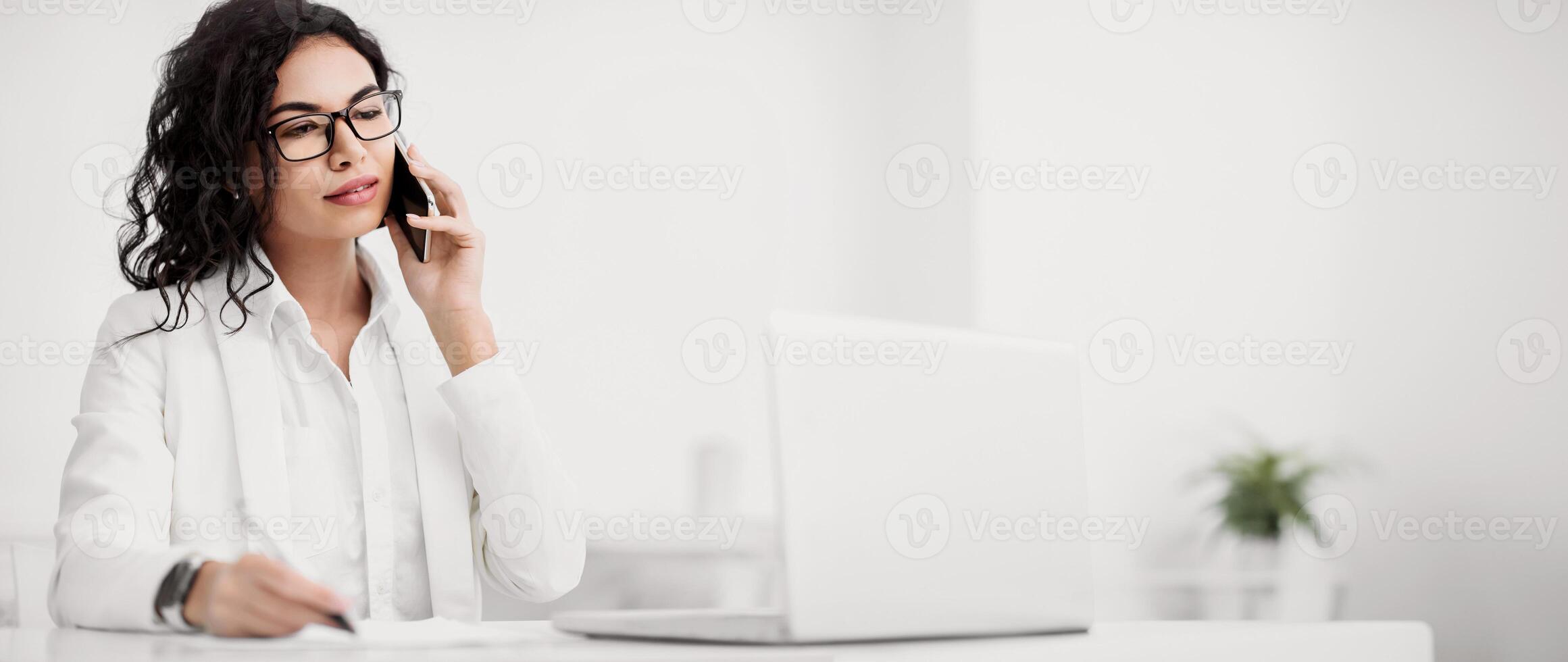 Business Call. Serious mexican woman talking on mobile phone, taking notes and working on laptop. Copy space photo