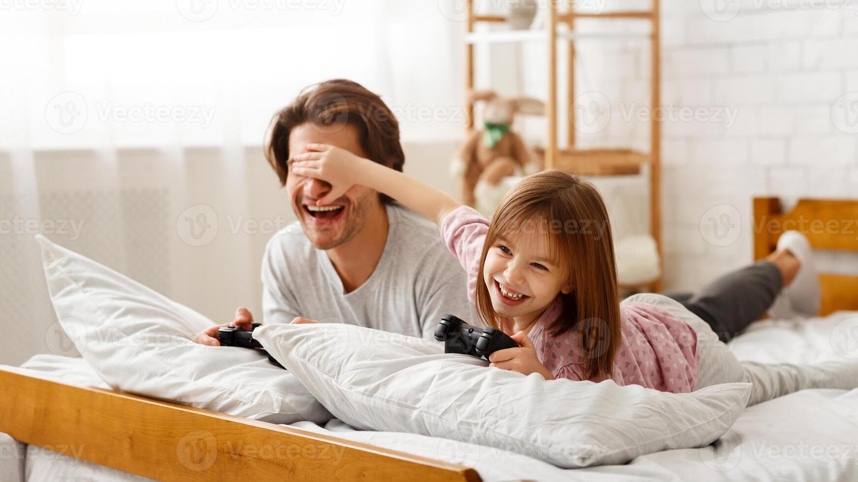 Playful kid girl covering her dad eyes while playing videogame, cheating to win, free space photo