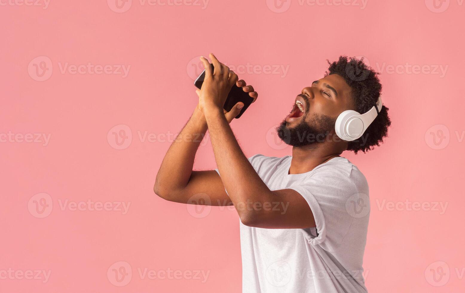 Emotional african american man dancing, singing his favorite song on smartphone with closed eyes, using phone as microphone, pink studio background photo