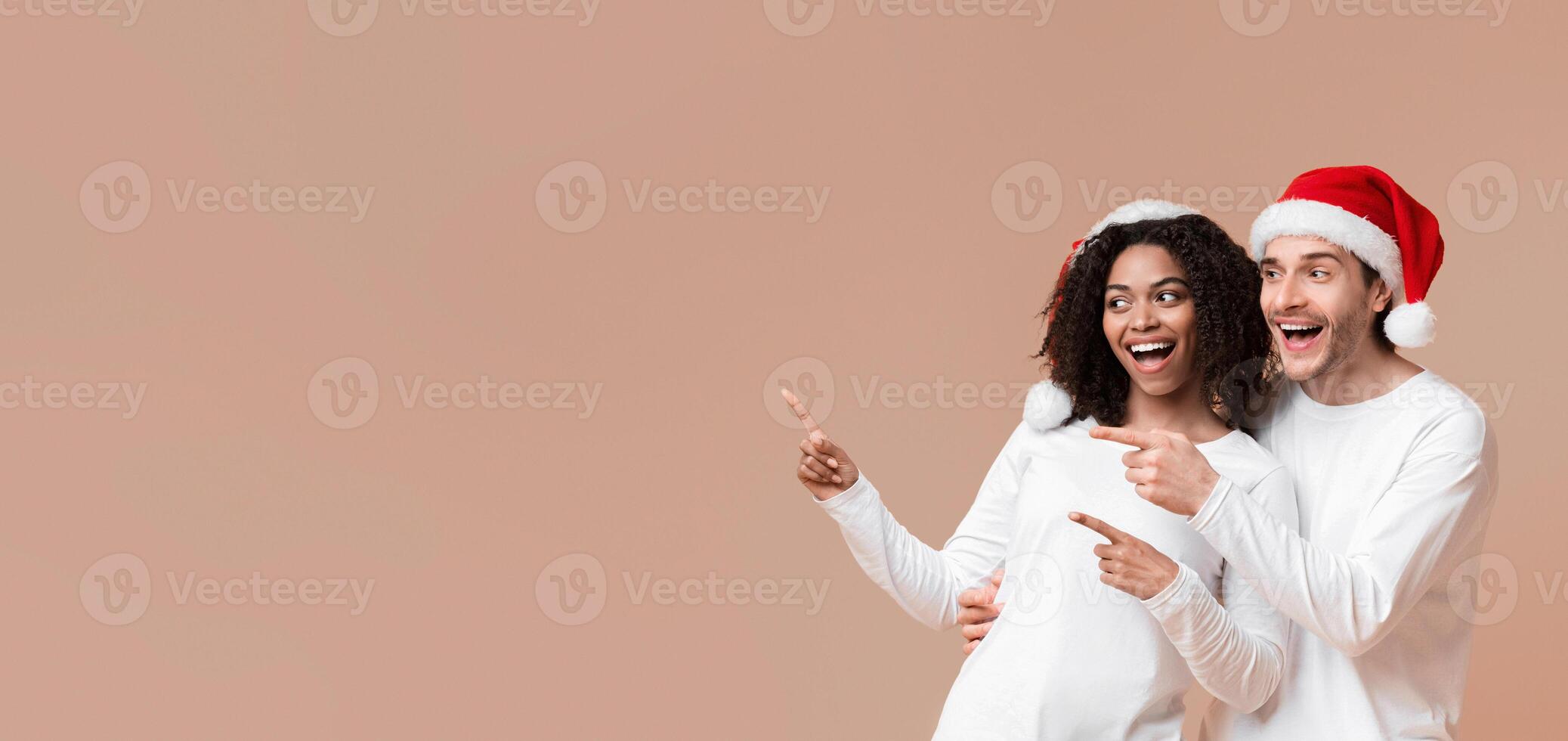 Happy interracial spouses in santa hats pointing with fingers at copy space on yellow background, demonstrating place for your ad, panorama photo