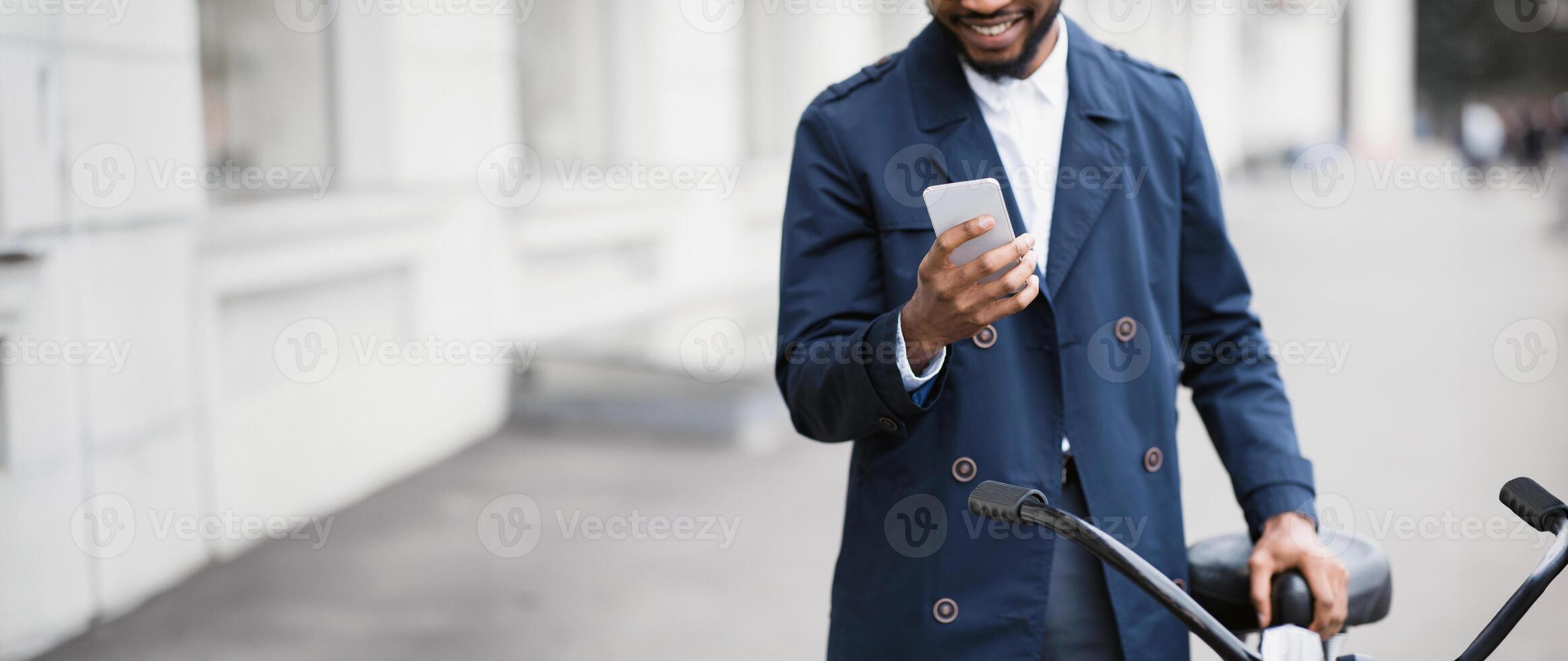 An African American businessman is commuting to his office by bicycle, using his phone. The mood is dynamic and contemporary, emphasizing multitasking and active transportation, cropped photo