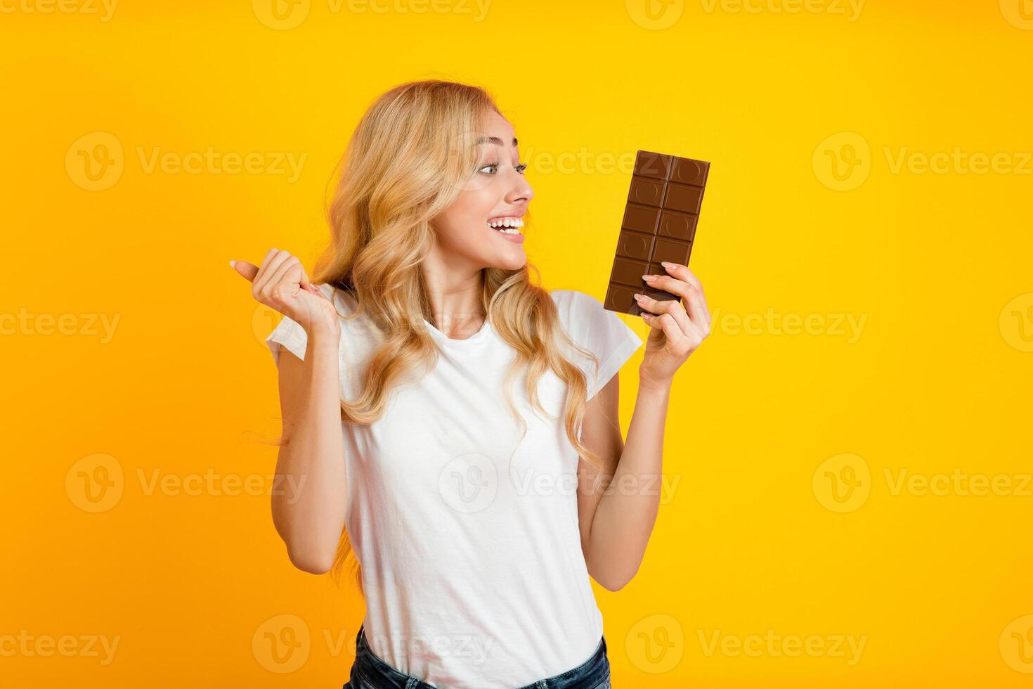 A young blonde woman with a happy expression holds a chocolate bar in her right hand and points to the left side of the frame with her left hand photo