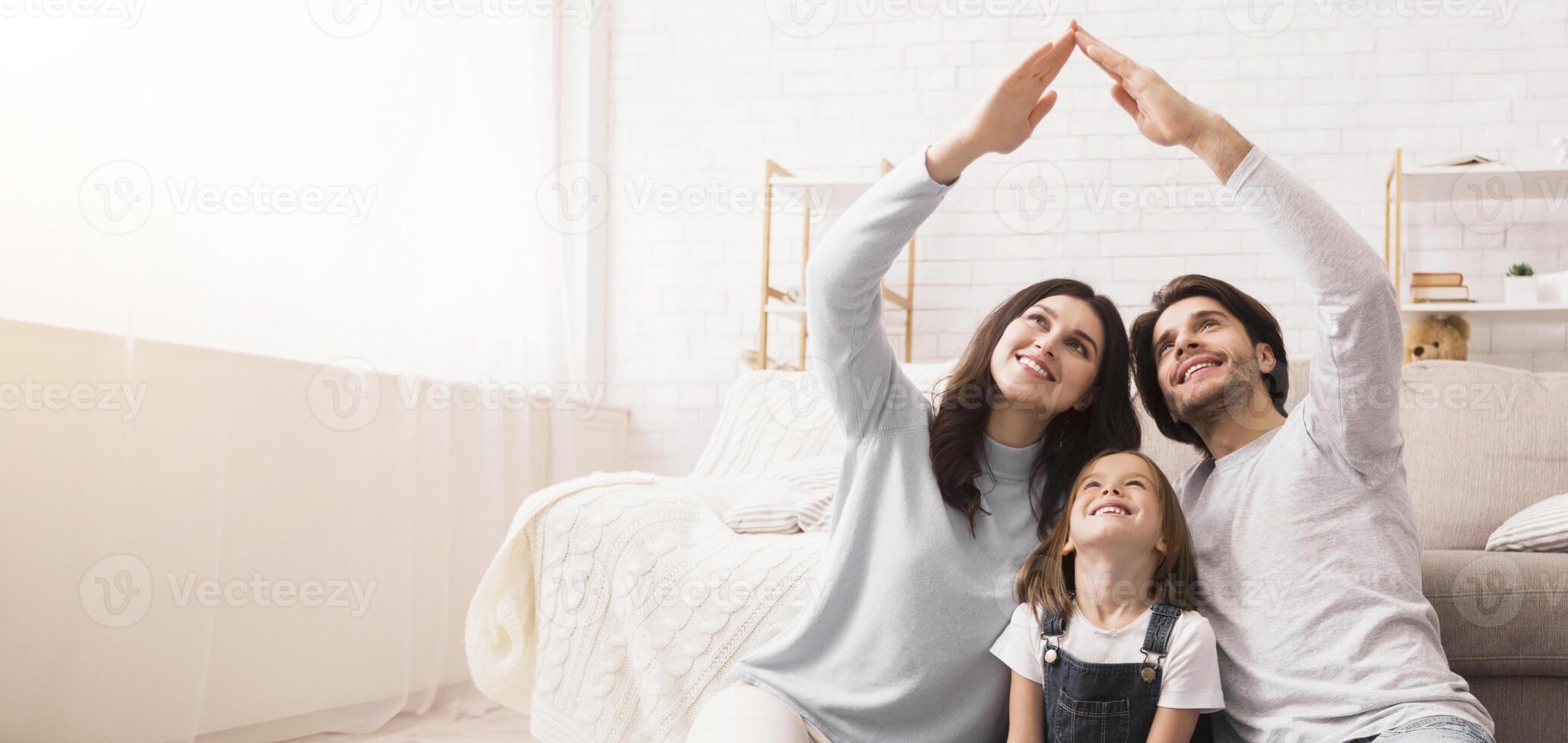 Family Love, Care And Protection. Happy Young Parents Making Symbolic Roof Of Hands Above Their Little Daughter's Head, Sitting Together On Floor photo