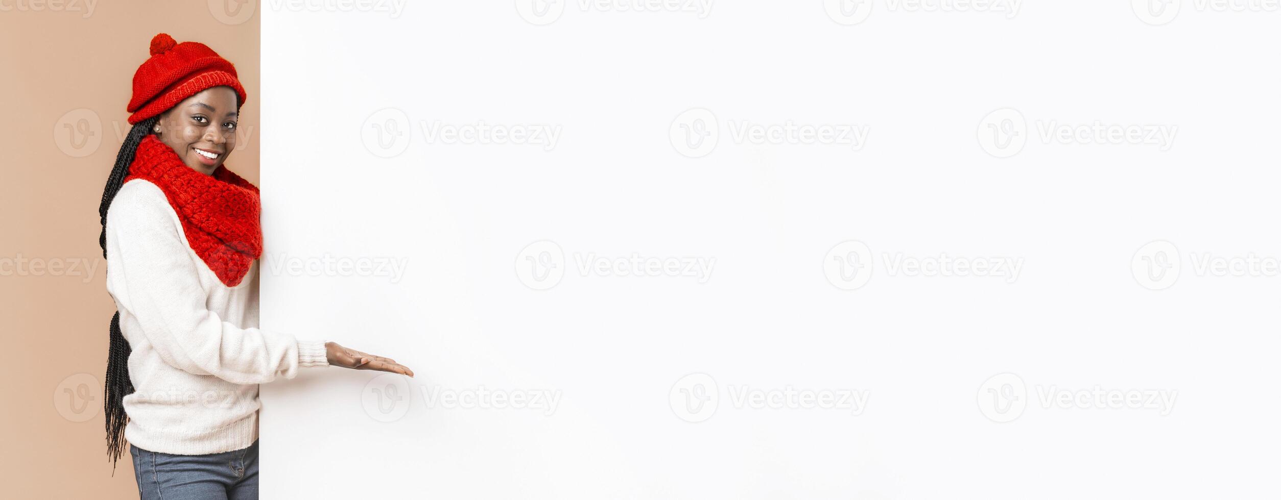 Empty Space For Advertising. Black cheerful woman in red knitted set directing with palm to empty white board, beige background, panorama photo