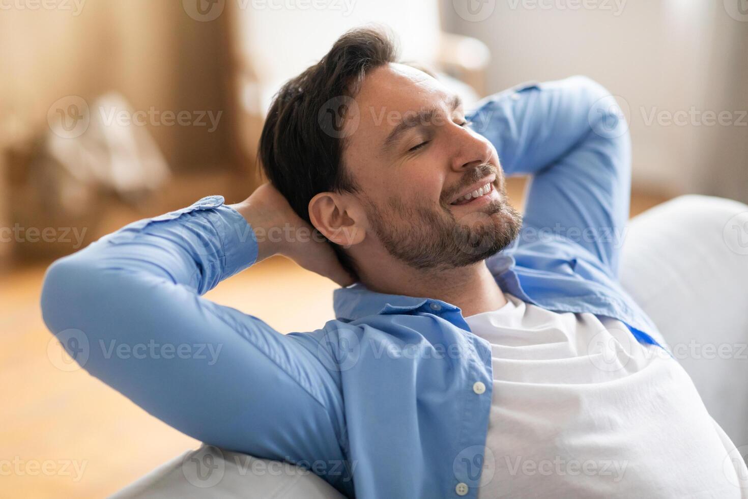 A man is seated on a couch, leaning back with his hands on his head. He appears relaxed and contemplative as he takes a moment to unwind at home. photo