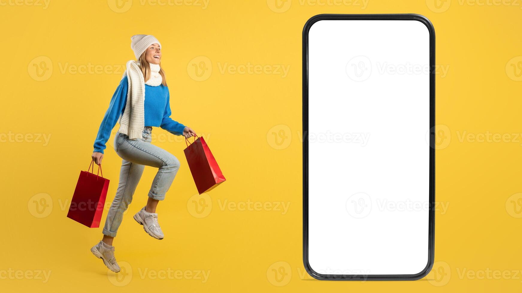 A joyful young woman in casual winter attire holds red shopping bags, leaping near a large smartphone screen with a blank display against a bright yellow backdrop. photo