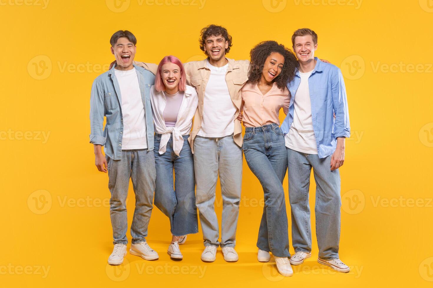 Five young adults are standing together in front of a yellow background. They are all smiling and laughing, and they are dressed in casual clothing. The group seems to be having a lot of fun photo