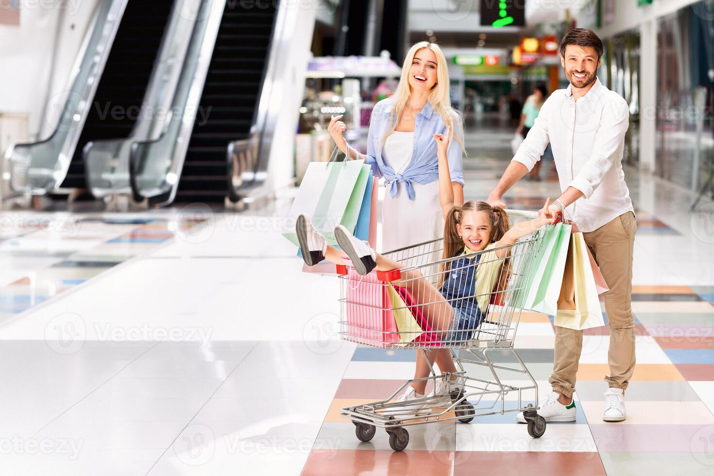 A young girl sits in a shopping cart being pushed by her father, while her mother walks behind with shopping bags. The family is smiling and laughing as they navigate the modern mall photo