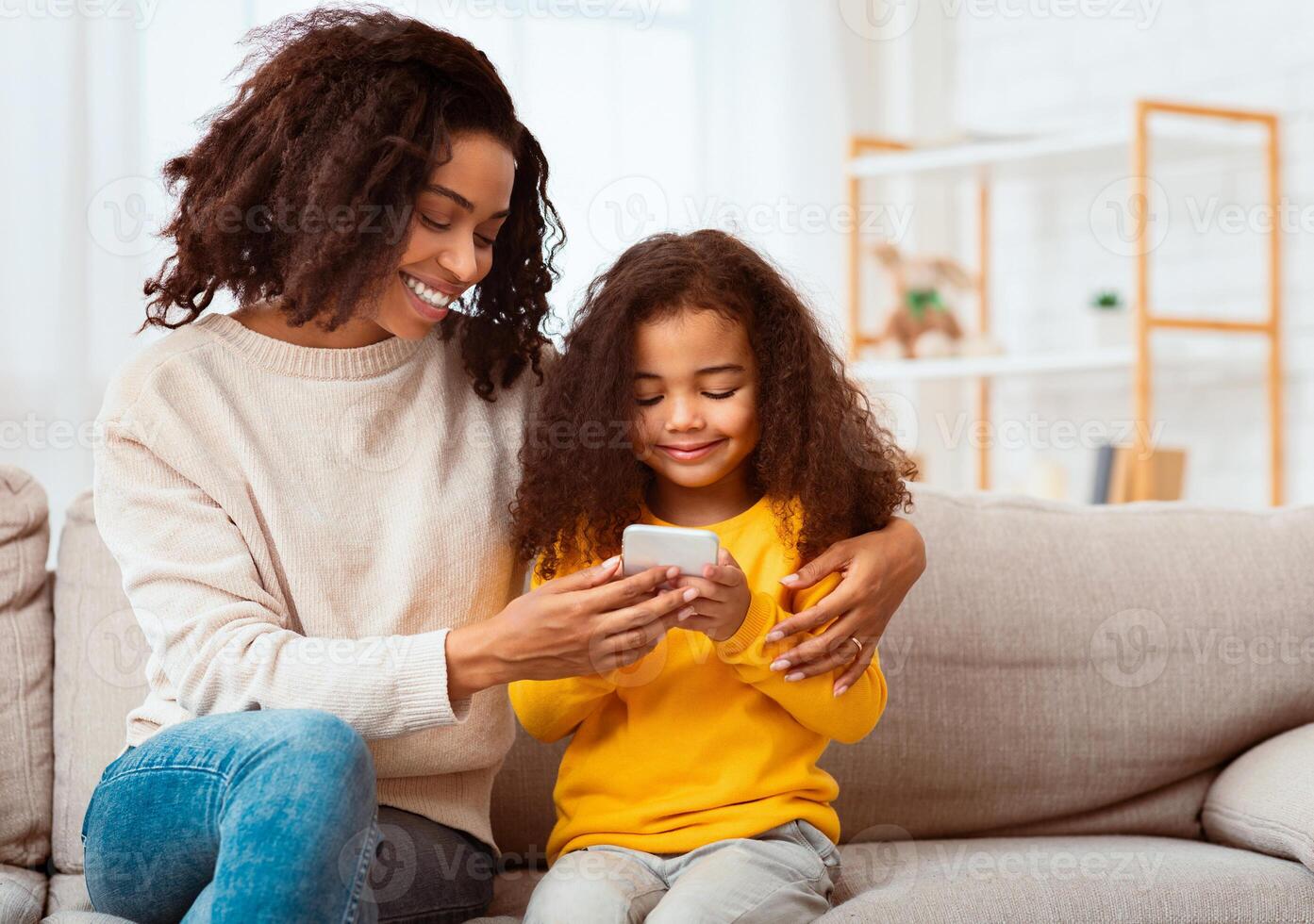 Family Weekend. Cheerful African American Mother And Daughter Using Smartphone Playing Mobile Games Together Sitting On Couch Indoor. photo