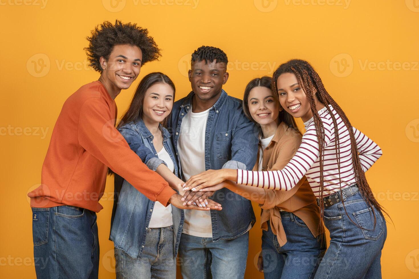 This image shows a diverse group of five friends stacking their hands together in a studio setting. They are all smiling and appear to be enjoying each others company. The background is bright yellow photo