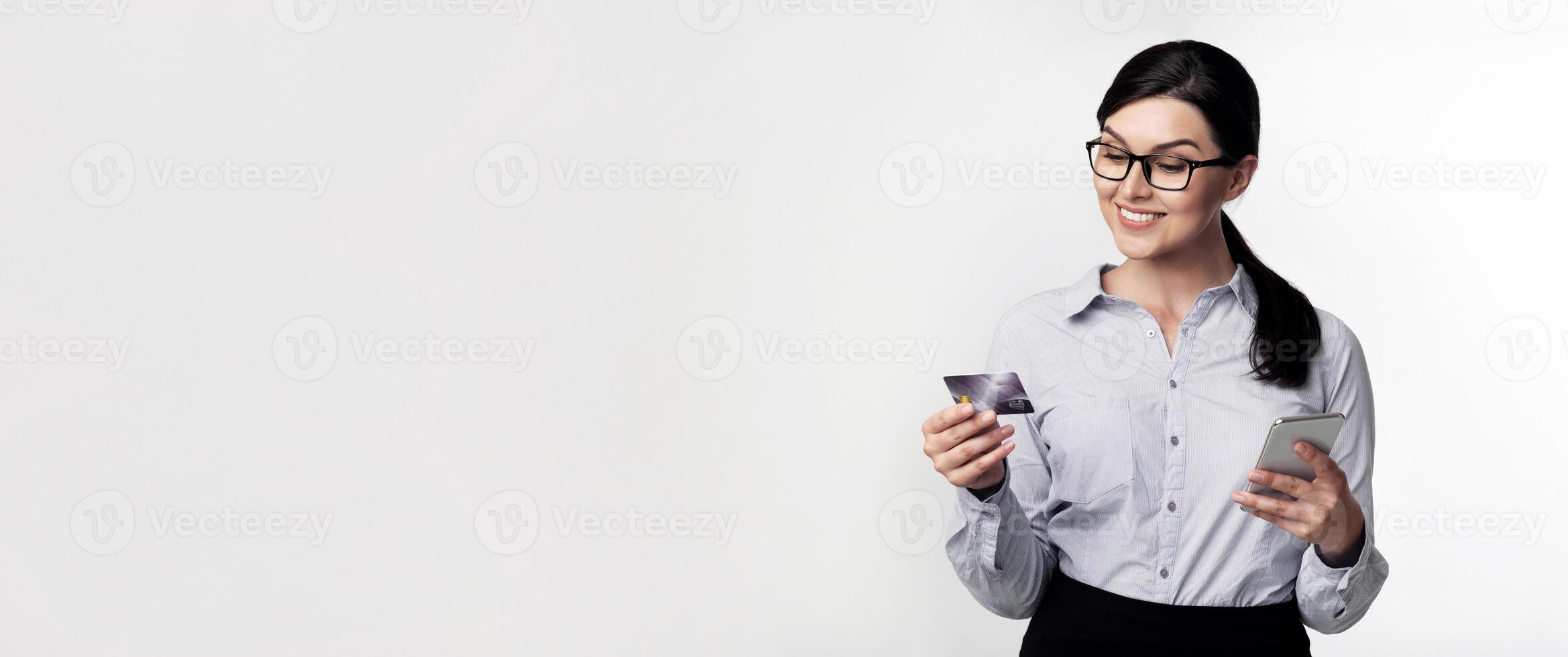 A woman wearing glasses and a white shirt is holding a credit card and a smartphone, smiling as she looks at the screen. photo