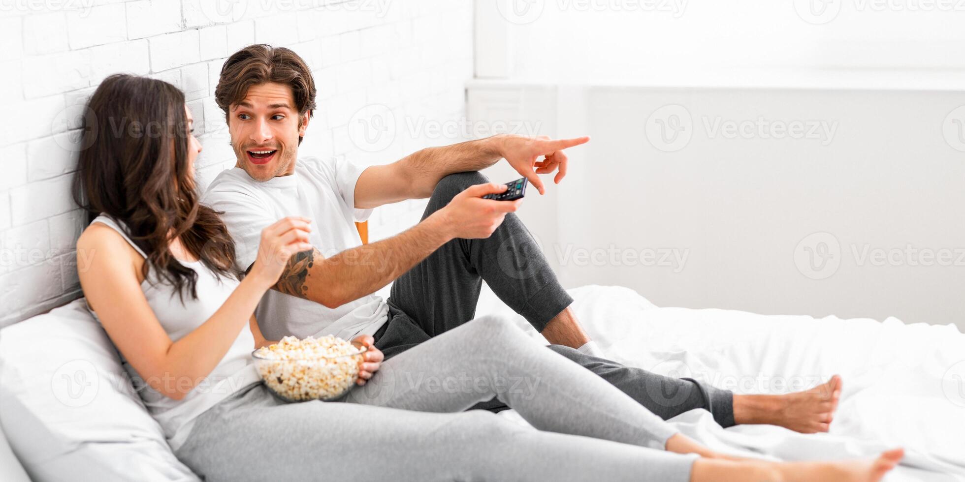 A young couple is comfortably sitting in bed together, watching television and enjoying a bowl of popcorn. The woman is smiling while the man is pointing at the TV and appears to be laughing. photo