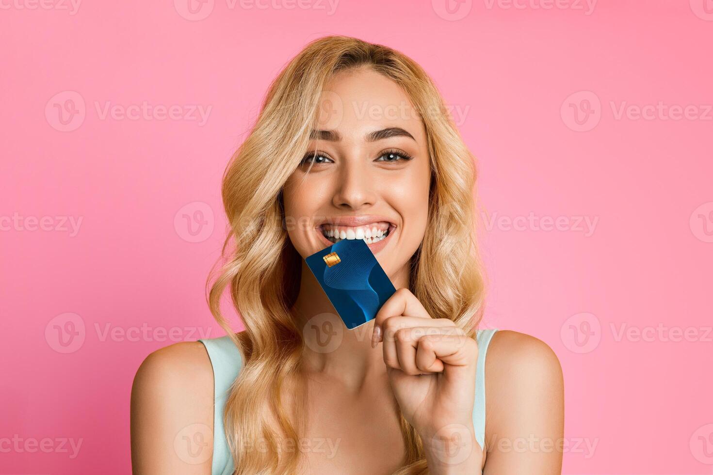 A smiling woman with long blonde hair is holding a credit card in front of her mouth. She is wearing a light blue tank top and is looking directly at the camera. The background is a solid pink color. photo