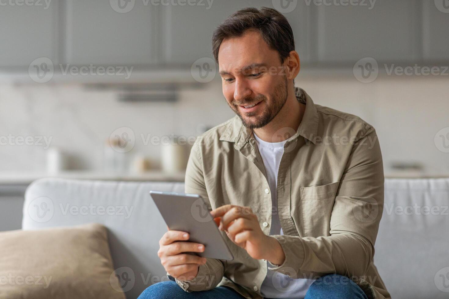 A man sits on a couch in a living room, smiling as he uses a tablet computer. He is wearing a beige button-down shirt and blue jeans. photo