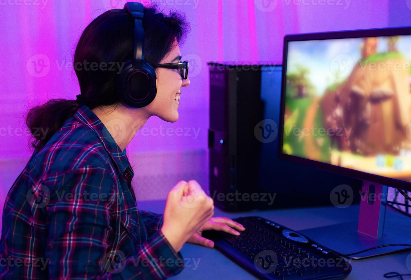 A young woman is pictured playing a game on a desktop computer. She is wearing a blue and red plaid shirt and headphones and is excitedly pumping her fist in the air. photo