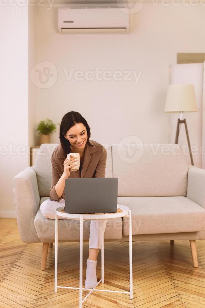 Surfing Internet. Happy smiling woman working on laptop computer, sitting on sofa, holding cup of coffee, copyspace photo