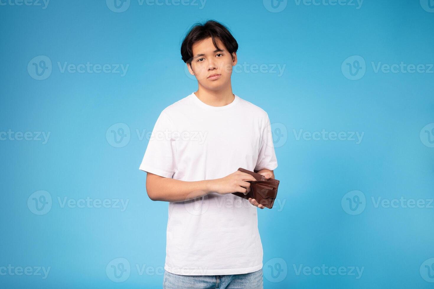 A teenage boy stands against a solid blue background, holding a brown wallet with a neutral expression. He is wearing a plain white t-shirt and jeans. photo