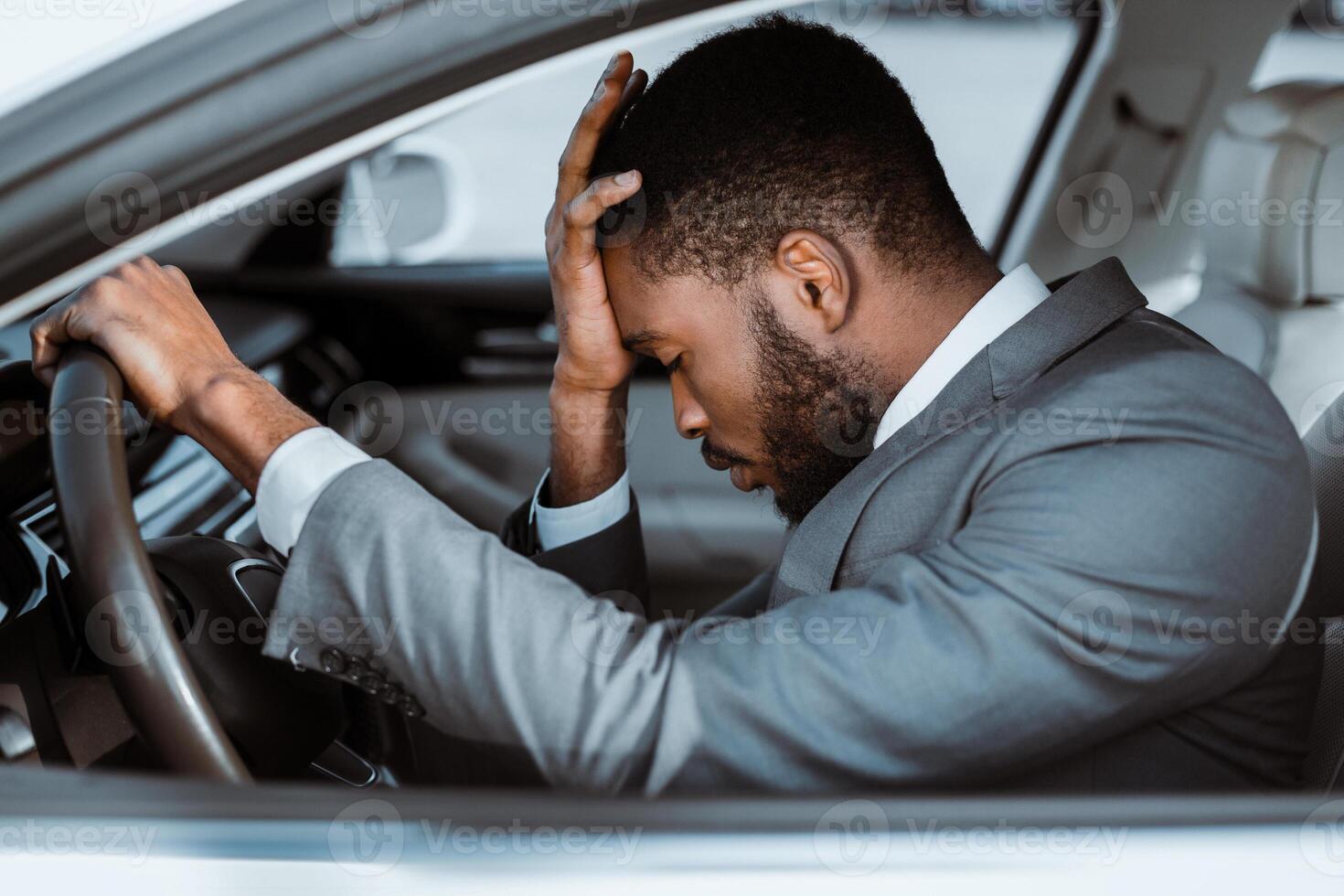 African American man in a grey suit is sitting inside the drivers seat of a car. He has his head in his hands, appearing stressed or frustrated. His hand rests on the steering wheel photo