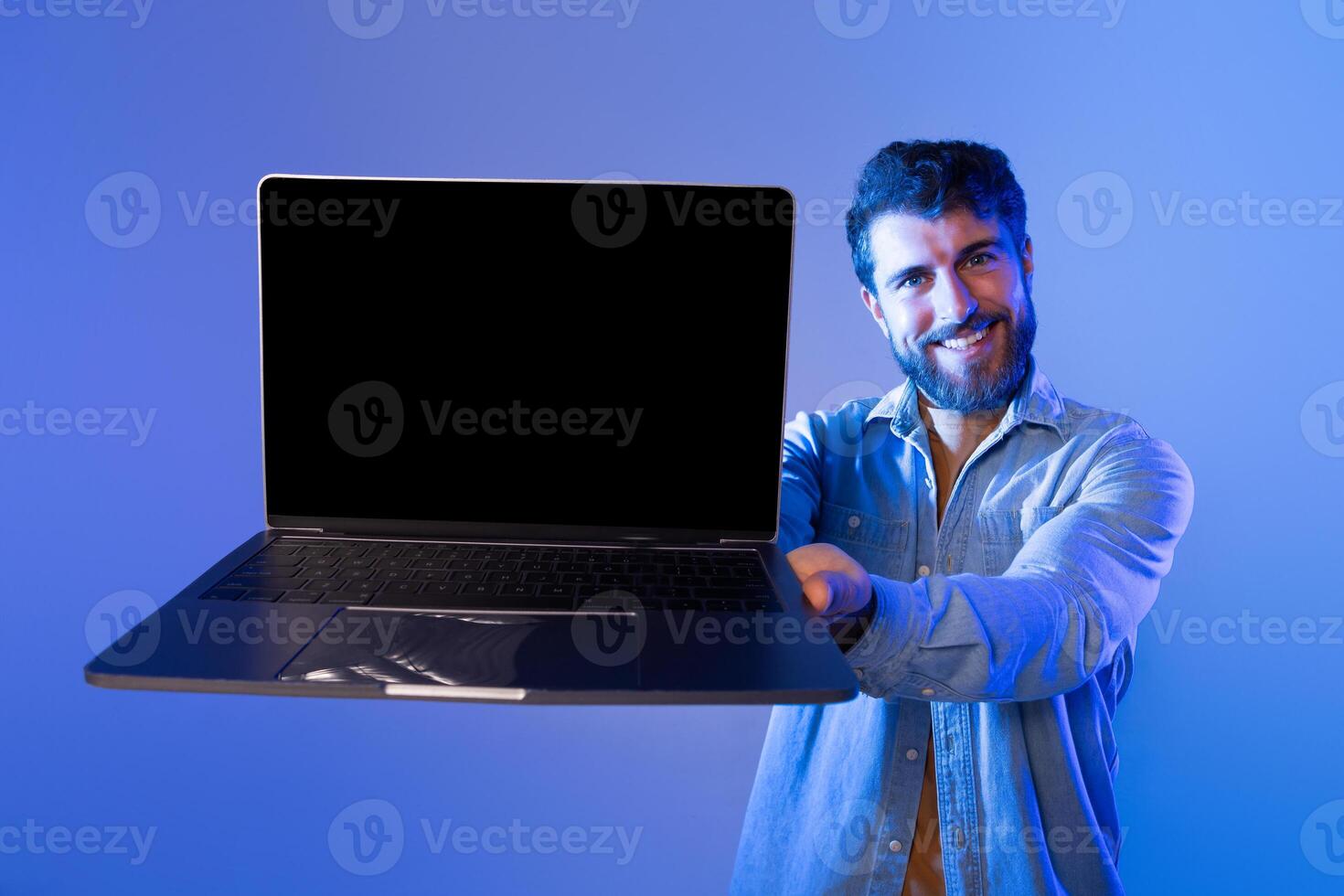 A cheerful man with a beard holds a laptop with a blank screen, showcasing it toward the camera. The setting features a blue gradient background and appears to be taken during the evening. photo