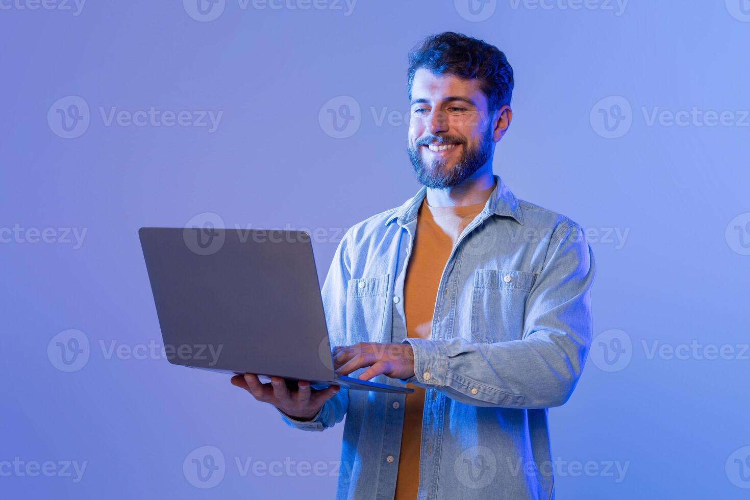 A man sitting at a desk is smiling while interacting with a laptop computer. He appears engaged and focused on the screen, possibly working or communicating with others online. photo