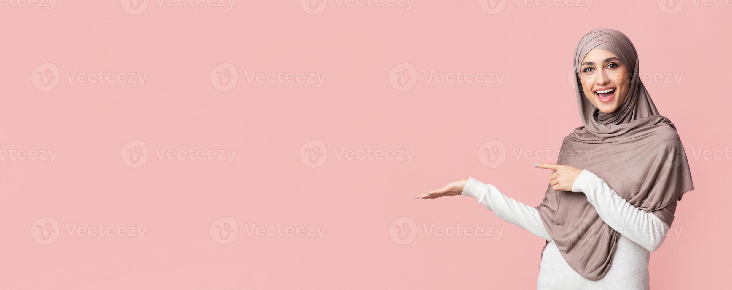 Beautiful muslim woman in hijab demonstrate something on her empty palm and pointing at it, standing over pink background, panorama with copy space photo