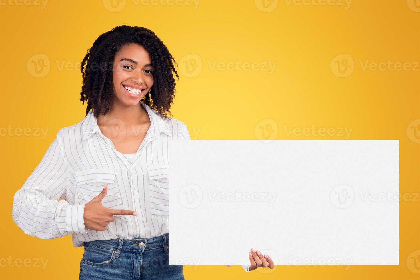 African Woman Holding Blank Paper On Yellow Background, Smiling And Pointing At It, Empty Space photo