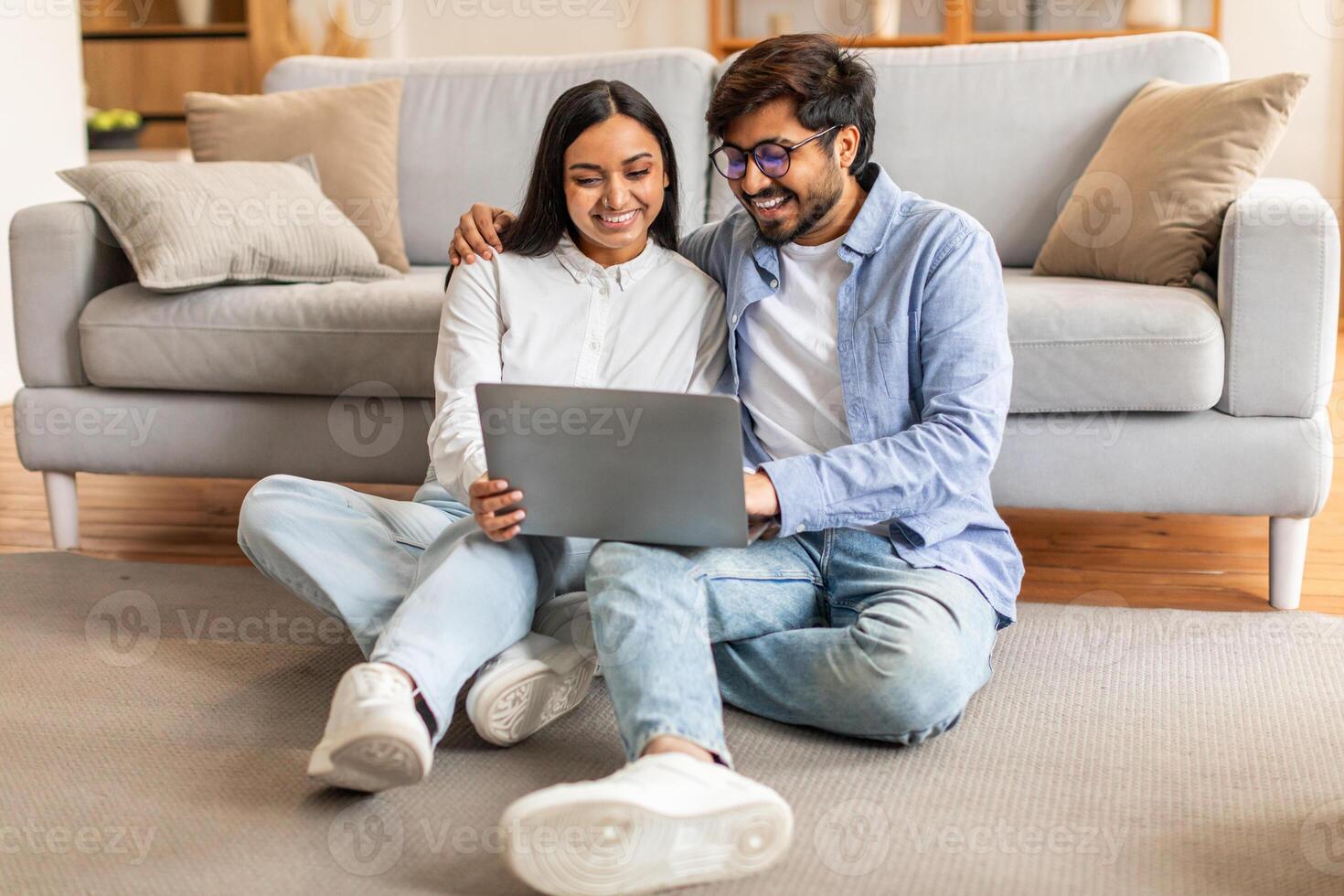 Indian man and woman are seated on the floor, focusing intently on a laptop screen in front of them. They appear engrossed in whatever they are viewing or discussing on the computer. photo
