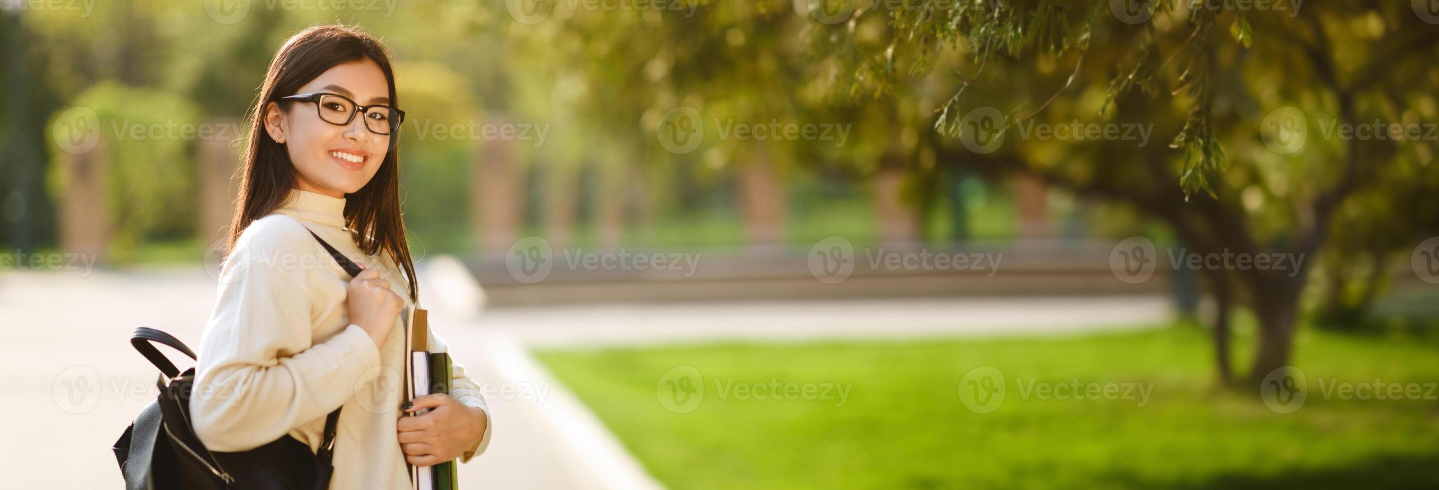 A young Chinese woman in a white sweater and glasses walks through a park. She is carrying a backpack and holding a stack of books in her left hand, panorama with copy space photo
