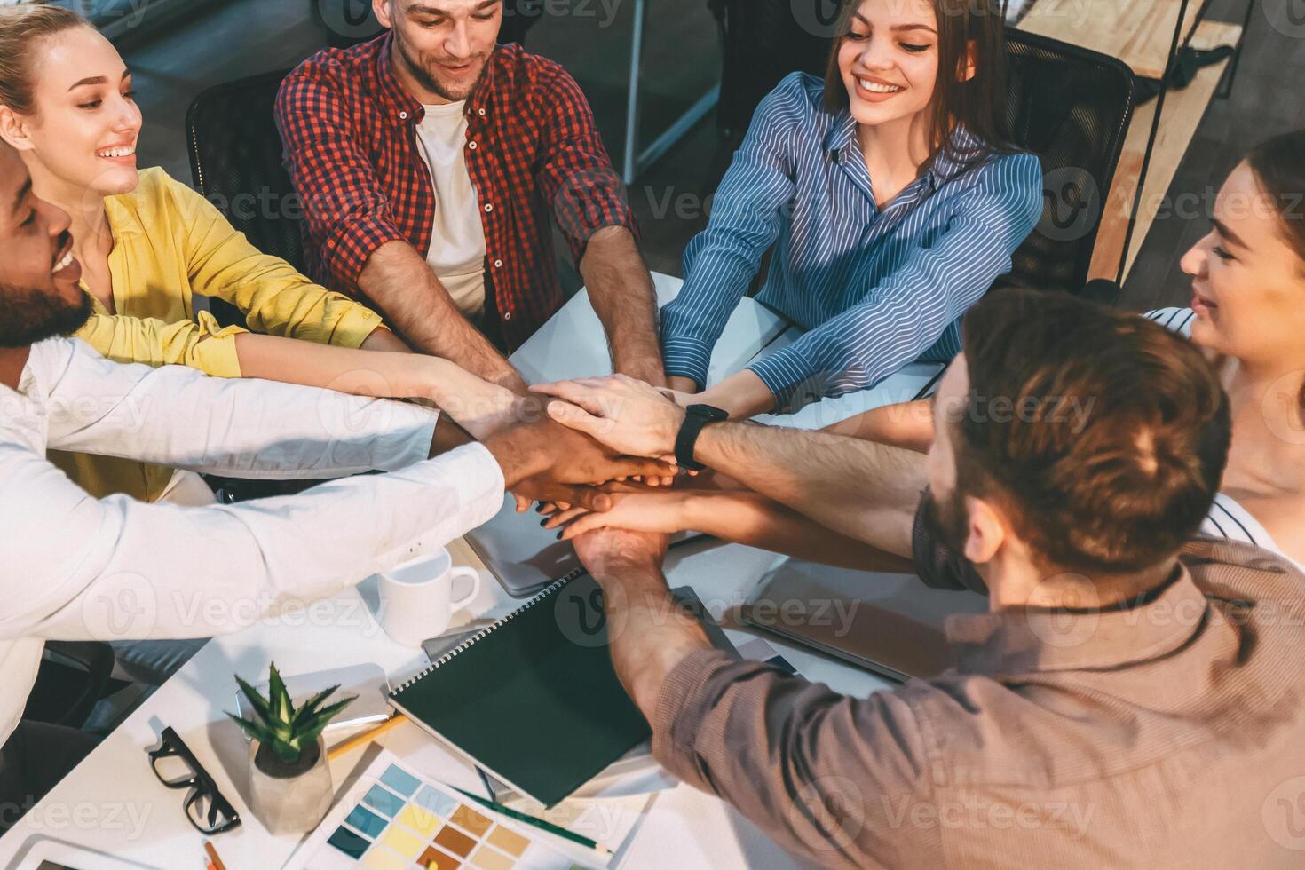 A group of diverse coworkers enthusiastically join hands over a desk, symbolizing teamwork and unity in a modern office environment. photo