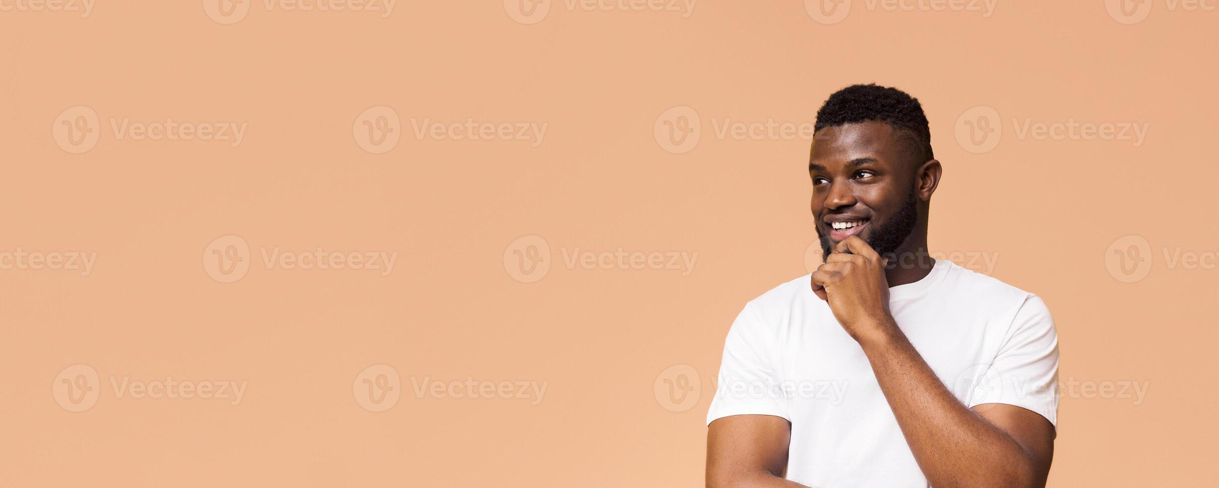 A young black man with a beard is smiling and looking off to the side with his hand on his chin. He is wearing a white t-shirt and is standing in front of a light brown background, copy space photo