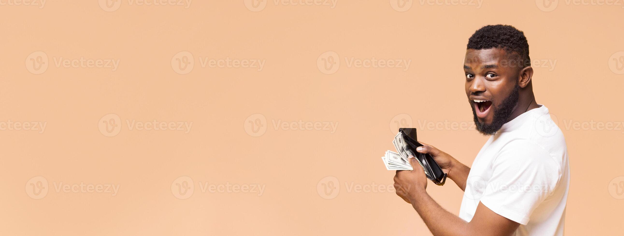 African American young man with a beard is seen holding a wallet and a wad of cash while looking towards the camera with a surprised expression, copy space photo
