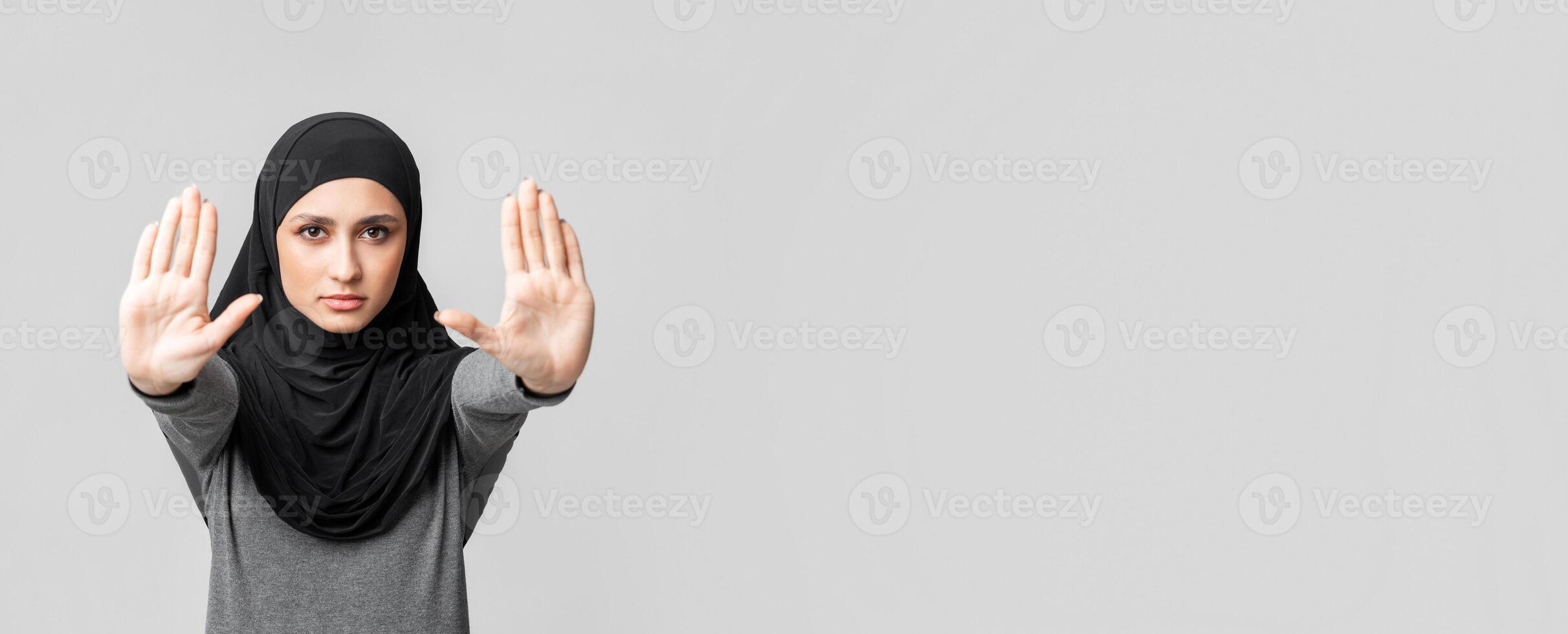 No discrimination. Concerned arabic woman in headscarf showing stop sign gesture with two hands, demonstrating palms over grey background with free space photo