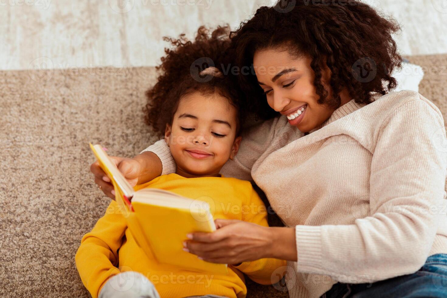Happy African American Mother And Daughter Reading Book Together Sitting On Sofa At Home. Family Weekend Leisure Concept photo