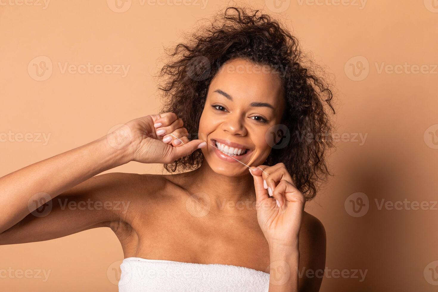 African American young woman with curly brown hair smiles as she uses floss to clean between her teeth. She is wearing a white towel and standing in front of a tan background photo