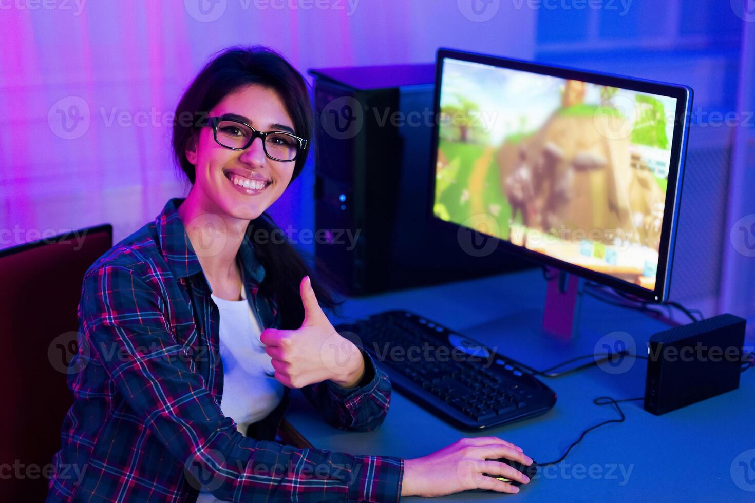 A woman wearing glasses smiles while playing a game on a computer. Her thumbs up gesture shows her enjoyment. She sits at a desk lit with purple and blue lighting in what appears to be a home photo
