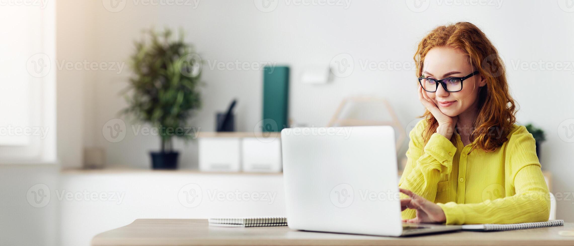 Remote Job Concept. Curly Red-Haired Woman In Specs Using Laptop Working At Home, Copy Space photo