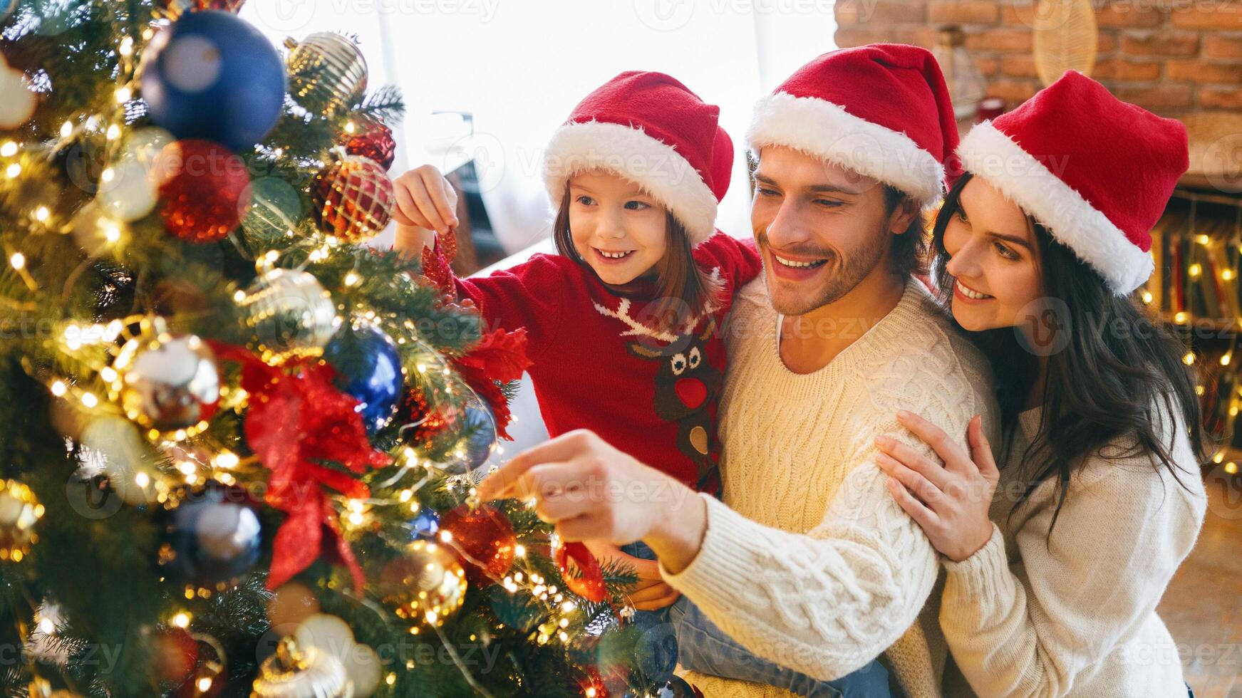 Happy family of three in Santa hats decorating Christmas tree with bubbles and bows at home photo