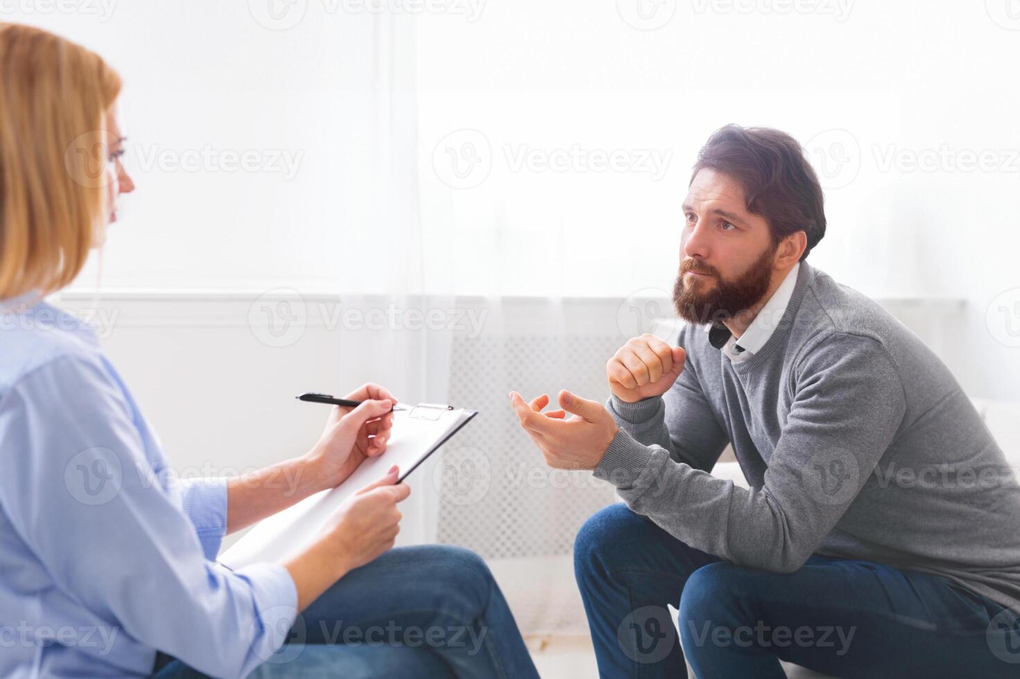 Handsome young man sitting on couch and talking to psychologist, doctor making notes, panorama with empty space photo