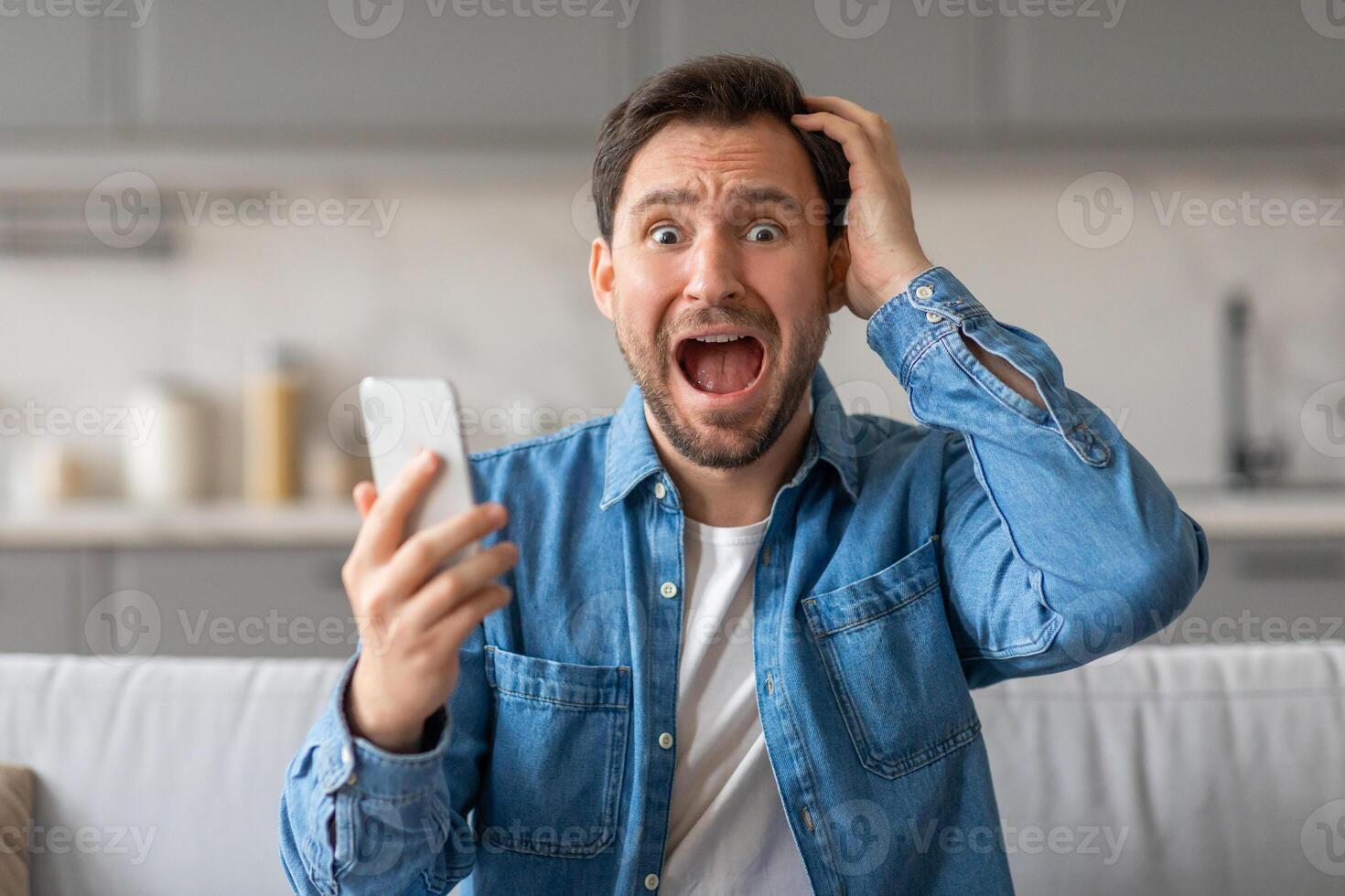 A man in a blue button-down shirt sits on a couch, looking shocked while holding a smartphone in his left hand and holding his right hand to his head. His mouth is open in surprise. photo