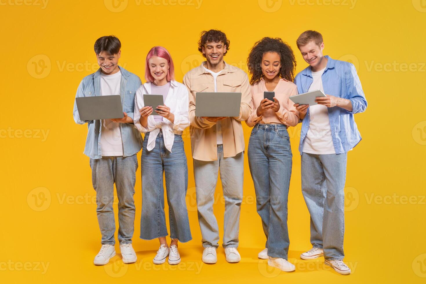 Five young adults, standing in a row in front of a yellow background, are using different forms of technology, including laptops, tablets, and smartphones. photo