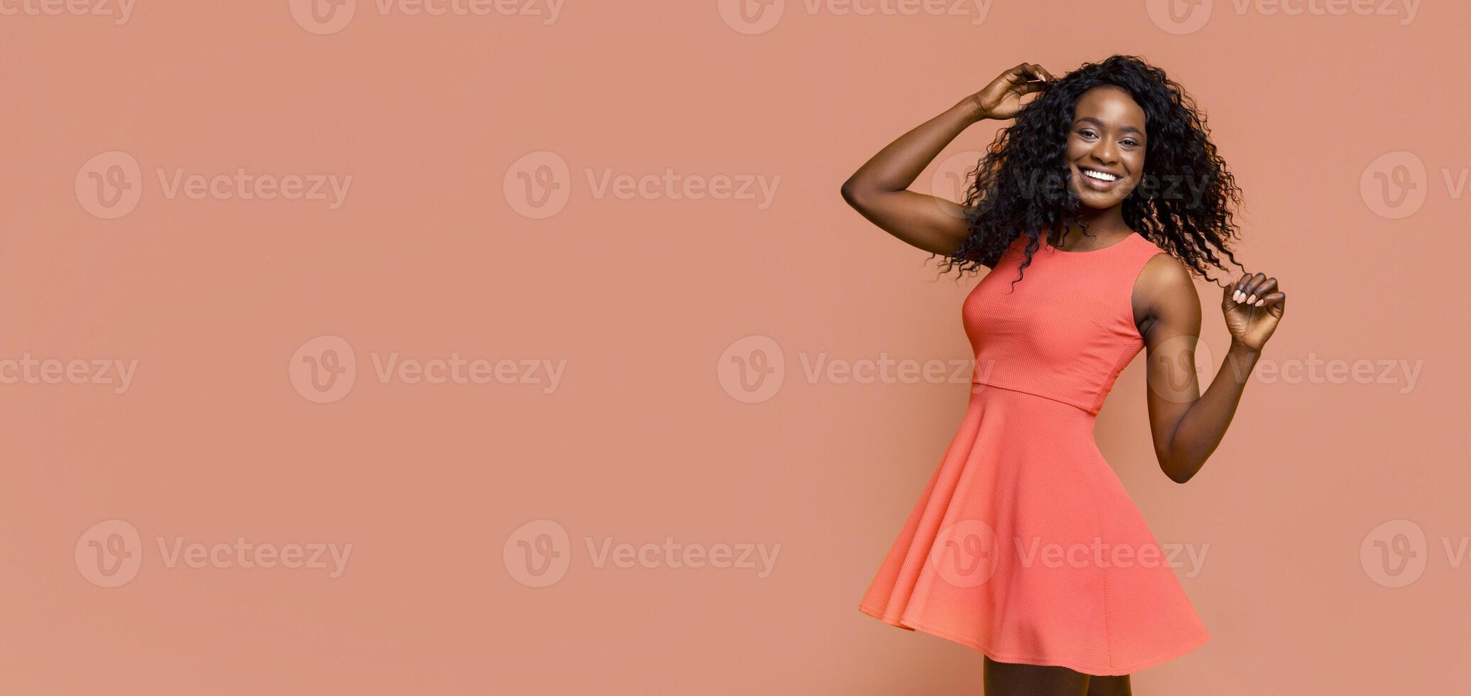 A Black woman with curly hair is smiling and posing in a coral colored dress. She is standing in front of a peach background. She is wearing a light pink shade of lipstick. photo