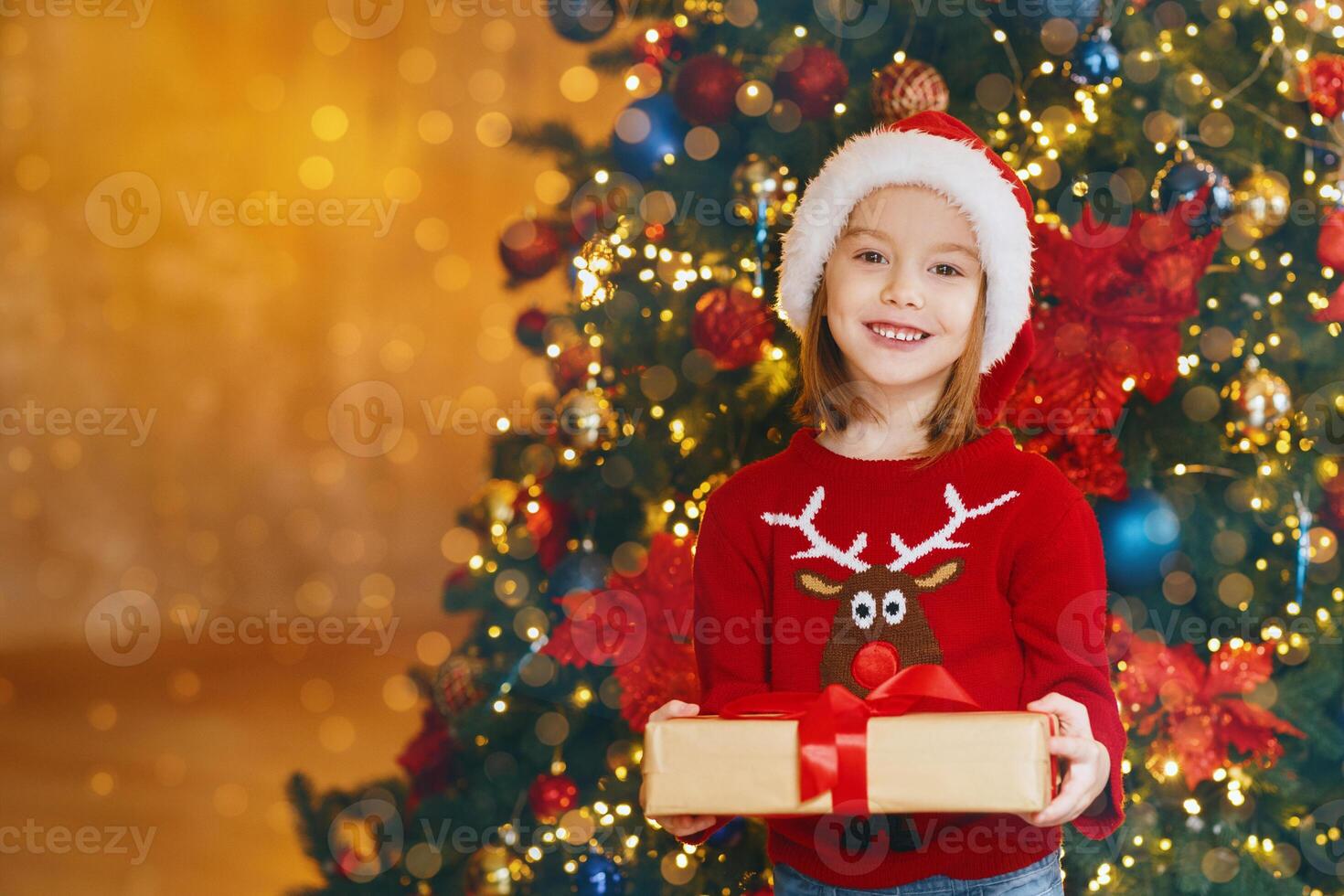 Little santa girl standing by xmas tree holding Christmas gift, smiling at camera, copy space photo