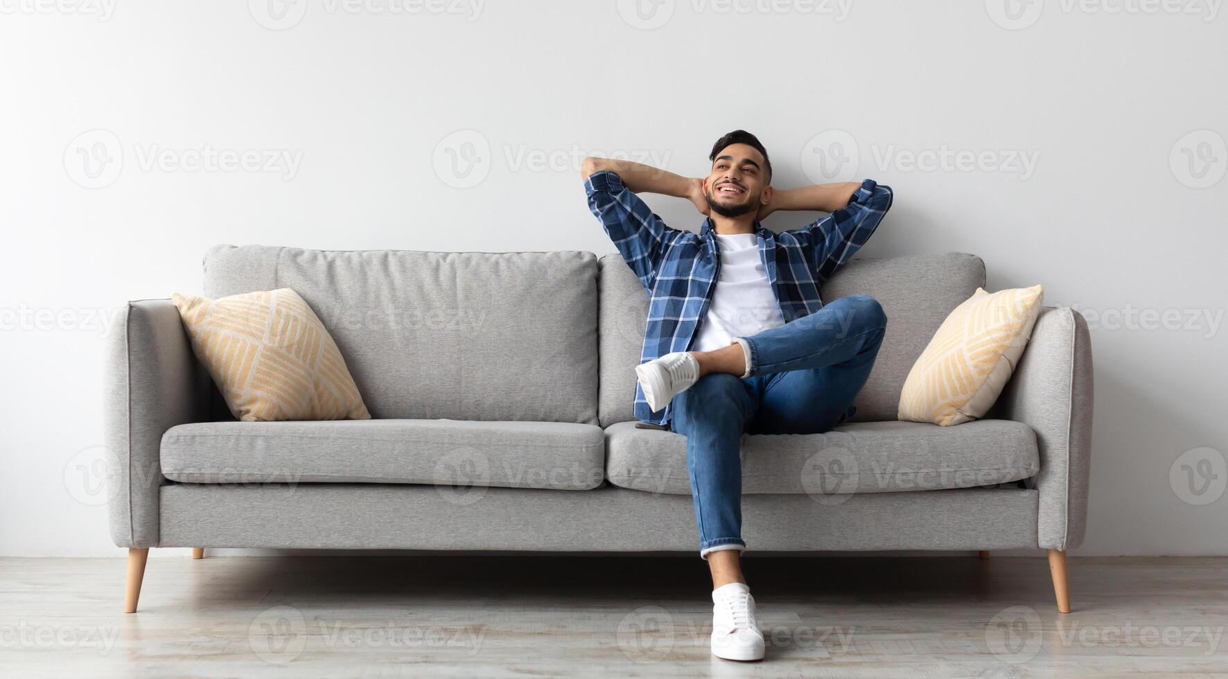 Rest Concept. Happy Middle Eastern guy sitting on comfortable couch at home in living room. Cheerful casual man relaxing on sofa, leaning back, enjoying weekend free time or break from work photo