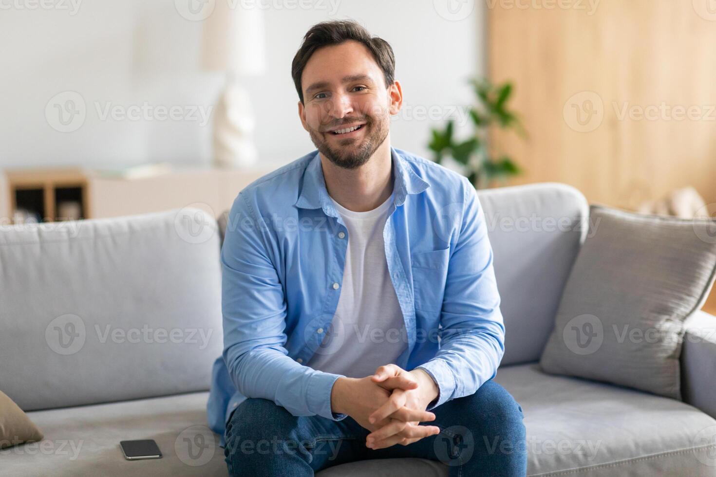 A man is seated on a beige couch in a cozy living room. The room features a coffee table, a lamp, and a bookshelf. The man appears relaxed and is looking at his phone. photo